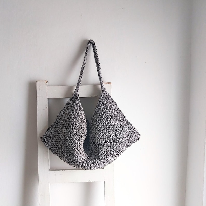 May include: A gray crocheted tote bag with a wide, open top and two long handles. The bag is hanging on a white wooden ladder.