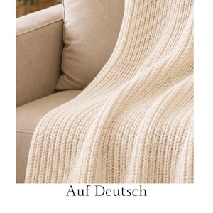 May include: A cream-colored knitted throw blanket with a prominent vertical ribbed texture. The fabric features a consistent chunky knit pattern that drapes over the side of a matching cream-colored upholstered furniture piece with a smooth finish.
