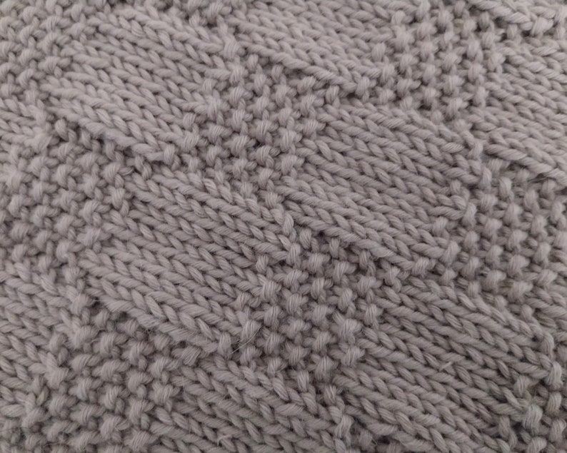 May include: Close-up of a gray knitted fabric with a textured pattern. The knit has a combination of raised and flat stitches, creating a visually interesting surface. The color is a muted, neutral gray.