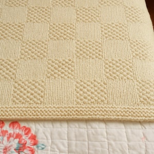 May include: Cream-colored knitted blanket with a checkerboard pattern. The blanket features alternating squares of textured and ribbed knit stitches. The bottom edge has a knitted border. The blanket is resting on a floral patterned bedspread.