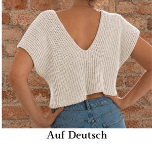 May include: A cream-colored, short-sleeved knit top featuring a deep V-neckline on the back, a ribbed texture, and a cropped length, paired with blue denim jeans. The garment is constructed with a visible vertical knit pattern throughout the fabric.