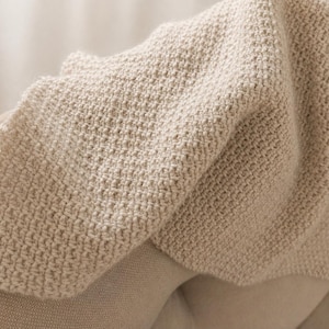 May include: A close-up of a beige knitted blanket draped over a light-colored surface. The blanket has a textured, woven appearance with visible stitches. The soft, neutral tones create a cozy and inviting aesthetic.
