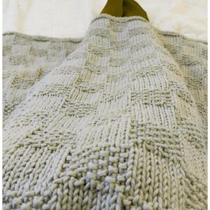 May include: A light gray knitted blanket with a textured, geometric pattern. The blanket is folded, revealing a glimpse of a green fabric on the top. The knitted design features a repeating square pattern.