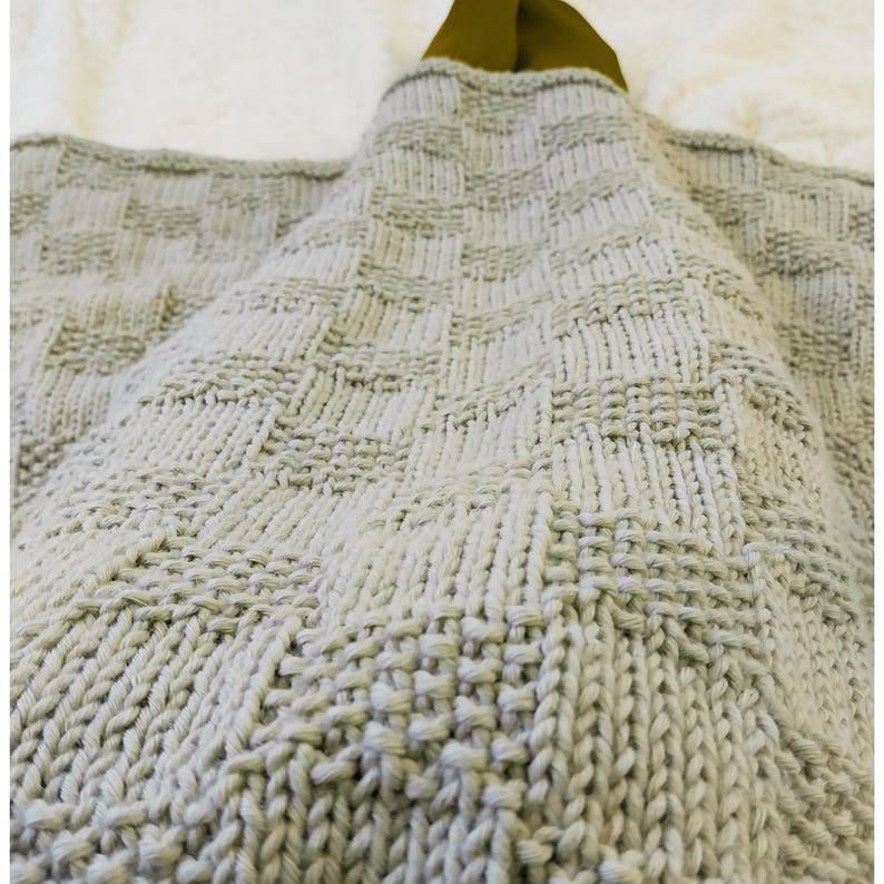 May include: A light gray knitted blanket with a textured, geometric pattern. The blanket is folded, revealing a glimpse of a green fabric on the top. The knitted design features a repeating square pattern.