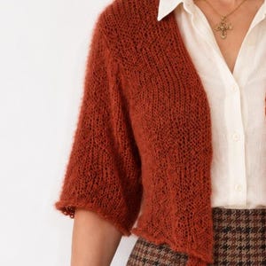 May include: A rust-colored, open-front cardigan featuring a loose, airy knit texture with three-quarter length sleeves. The garment is worn over a white button-down shirt and a brown houndstooth patterned skirt, paired with a gold cross pendant necklace.