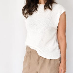 May include: A sleeveless white knit top with a round neckline and a straight hemline, paired with tan trousers. The top features a fine-gauge knit texture and a relaxed fit, while the trousers have a high-waisted design with a visible elastic waistband.