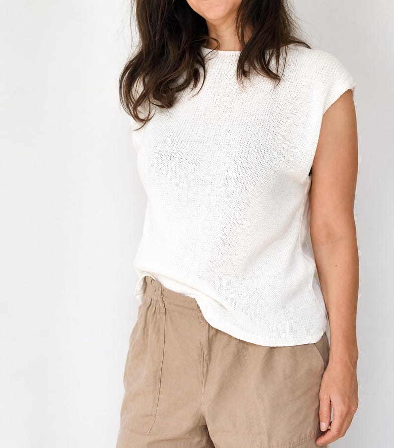 May include: A sleeveless white knit top with a round neckline and a straight hemline, paired with tan trousers. The top features a fine-gauge knit texture and a relaxed fit, while the trousers have a high-waisted design with a visible elastic waistband.