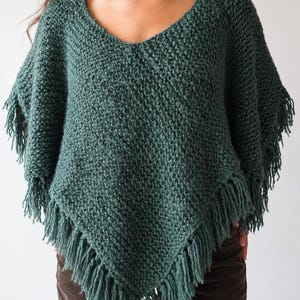 May include: A dark teal knitted poncho featuring a V-neckline and a textured stitch pattern. The garment has a pointed hemline finished with long, matching fringe detailing along the edges, providing a layered look over dark brown trousers.