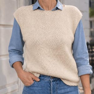 May include: A sleeveless beige knit sweater vest with a round neckline, worn over a light blue long-sleeved button-down shirt and blue denim jeans. The sweater vest features a simple stockinette stitch pattern and a relaxed fit.