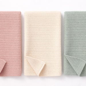 May include: Three folded crochet blankets in pastel shades. The blankets are in pink, cream, and sage green. Each blanket has a folded corner, revealing the textured stitch pattern. The blankets are neatly arranged, offering a cosy, handmade aesthetic.