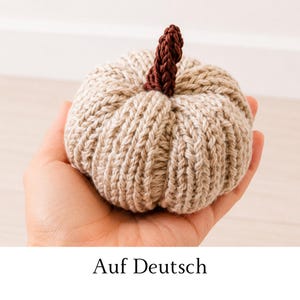 Könnte beinhalten: Ein kleiner, handgestrickter Kürbis in hellem Beige mit gerippter Textur. Der Kürbis hat oben einen dunkelbraunen, strukturierten Stiel aus Garn. Das Objekt ist kompakt und passt in die Handfläche.