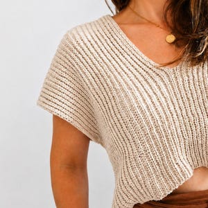 May include: A beige short-sleeved jumper featuring a prominent vertical ribbed knit texture and a V-neckline. The garment is cropped in length and constructed with a thick, textured yarn, showcasing a consistent pattern throughout the entire piece.