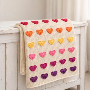 Op de afbeelding: Crèmekleurige gehaakte deken met rijen kleurrijke hartapplicaties. De harten zijn in tinten rood, oranje, geel, roze en paars. De deken is over een wit houten oppervlak gevouwen.