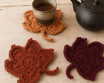 May include: Three crochet coasters shaped like autumn leaves in shades of orange, rust, and dark burgundy. One coaster supports a ceramic cup filled with tea, while a black cast iron teapot sits nearby, all featuring intricate yarn stitch patterns.