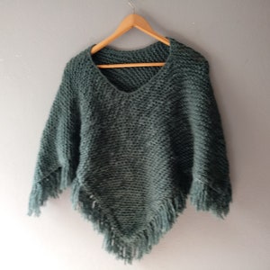 May include: A dark green knitted poncho with fringe detailing. The poncho is hanging on a wooden hanger against a light gray wall.