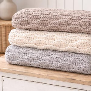 Könnte beinhalten: Drei gefaltete Strickdecken in neutralen Farben: Taupe, Creme und Grau. Jede Decke hat ein strukturiertes Zopfmuster. Die Decken sind auf einer weißen Holzoberfläche gestapelt, was auf ein gemütliches Wohn-Thema hindeutet.