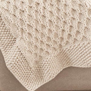 Puede incluir: Una manta de punto color crema que presenta un patrón central de rombos con trenzas, bordeado por un borde texturizado en punto de arroz. La construcción de hilo grueso crea una superficie táctil en relieve con un tono blanco roto uniforme.