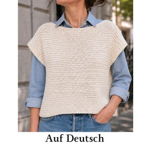 May include: A cream-colored sleeveless knit sweater vest with a horizontal ribbed texture, worn over a light blue collared button-down shirt and blue denim jeans. The sweater features a wide neckline and a boxy, cropped silhouette.
