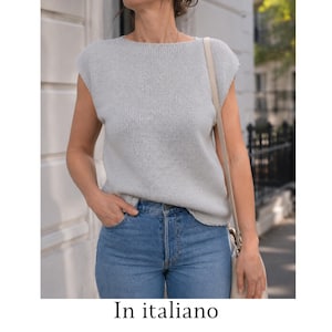 May include: A light gray sleeveless knit top with a wide boat neckline and a straight hem, paired with blue denim jeans. The top features a simple stockinette stitch pattern and a relaxed fit, worn with a beige shoulder bag strap visible on the side.