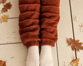 May include: A pair of rust-colored knitted leg warmers with a ribbed texture, worn over plain white knitted socks. The leg warmers feature horizontal ridges and extend from the lower calf down to the ankle, covering the top of the white socks.