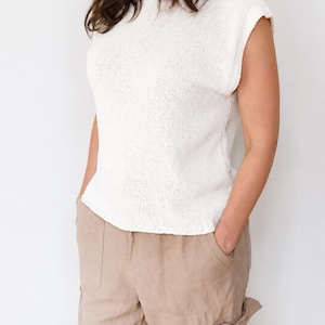May include: A sleeveless white knit top with a round neckline and a textured stitch pattern, paired with light tan cargo-style trousers featuring side pockets and a relaxed fit. The top has a straight hemline and a simple, clean construction.
