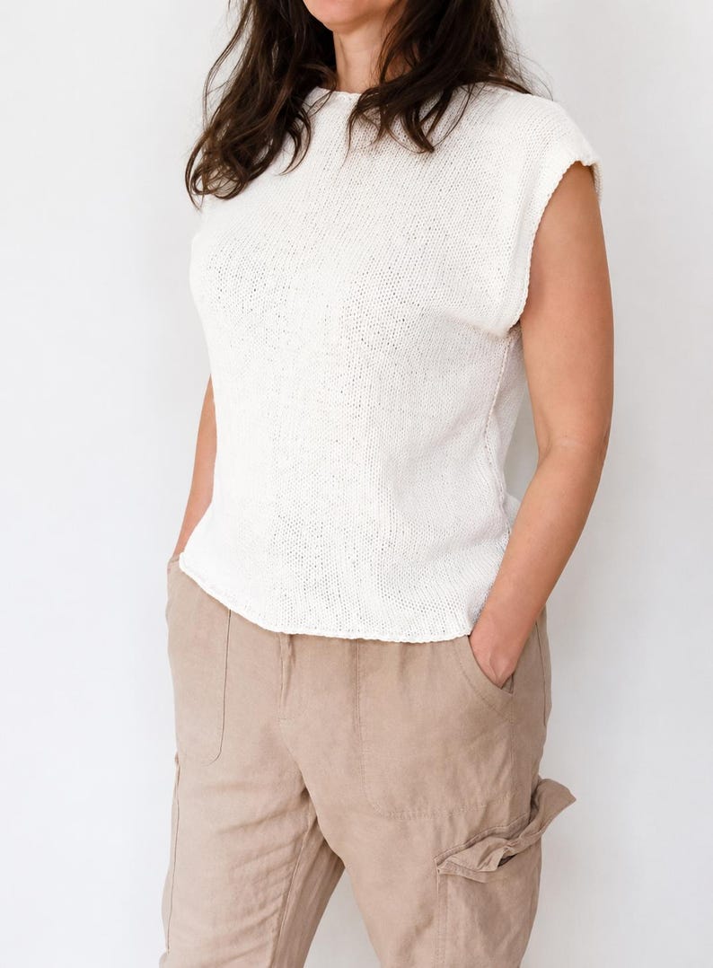 May include: A sleeveless white knit top with a round neckline and a textured stitch pattern, paired with light tan cargo-style trousers featuring side pockets and a relaxed fit. The top has a straight hemline and a simple, clean construction.