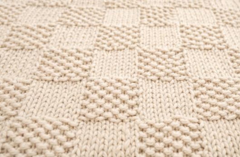 May include: Close-up of a cream-colored knitted blanket with a textured, geometric pattern. The design features alternating squares and diagonal rows of raised stitches, creating a visually appealing and tactile surface.