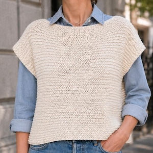 Peut inclure: Un gilet en tricot sans manches de couleur crème avec une large encolure bateau et une texture côtelée horizontale, porté sur une chemise boutonnée bleu clair à manches longues et un jean bleu avec une poche et un bouton en métal.