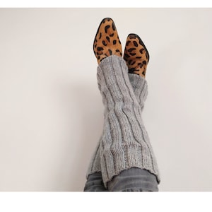 May include: Gray knitted leg warmers with a cable knit pattern. The leg warmers are worn over a pair of jeans and leopard print shoes.