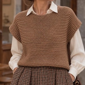 May include: A brown sleeveless knit sweater vest with a garter stitch texture, worn over a white long-sleeved collared shirt and a brown and black plaid skirt. The sweater vest features a wide round neckline and broad shoulder coverage.