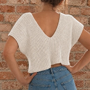 May include: A cream-colored, short-sleeved knit top featuring a deep V-neckline on the back, a ribbed texture, and a cropped hemline, paired with blue denim jeans. The garment is constructed with a vertical knit pattern and wide sleeves.