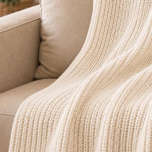 Puede incluir: Una manta de punto de color crema que presenta una textura acanalada vertical prominente. El tejido muestra un patrón de punto grueso y consistente con canales profundos y definidos que recorren toda la longitud del textil, creando una apariencia suave.