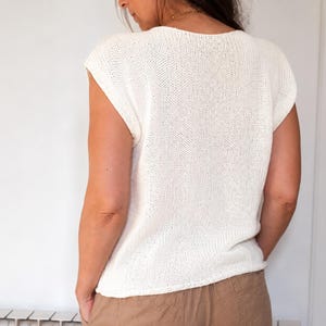 May include: A sleeveless, off-white knitted top with a wide, rounded neckline and a textured, open-stitch pattern. The garment features a relaxed fit with cap sleeves and a straight hemline, crafted from a lightweight, breathable knit fabric.