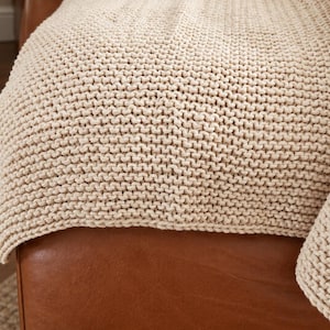 May include: A cream-coloured knitted throw blanket featuring a consistent garter stitch pattern. The textile has a thick, textured appearance with visible loops throughout the fabric, draped over a smooth, brown leather surface.