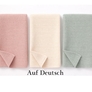 May include: Three folded blankets in soft colors: pink, cream, and green. The blankets have a textured, woven appearance. The text "Auf Deutsch" is printed in black at the bottom of the image.