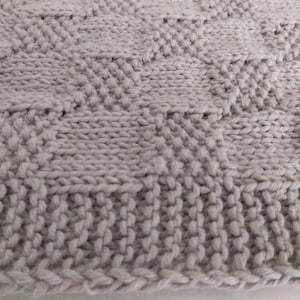 May include: Close-up of a light gray knitted blanket. The blanket features a textured pattern with a combination of vertical and diamond-shaped knit stitches. The edges are finished with a ribbed border.