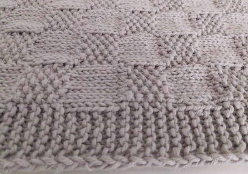 May include: Close-up of a light gray knitted blanket. The blanket features a textured pattern with a combination of vertical and diamond-shaped knit stitches. The edges are finished with a ribbed border.