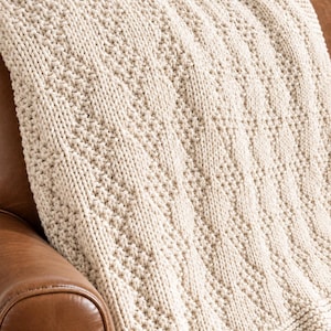 Pode incluir: Uma manta de malha na cor creme com um padrão de diamante texturizado criado através de pontos de meia e tricô alternados. A construção em fio grosso exibe um design geométrico consistente e repetitivo em toda a superfície.