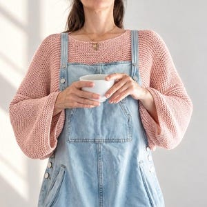 May include: A light pink long-sleeved knit jumper worn under light blue denim dungarees. The jumper features a loose, textured weave and wide sleeves. The denim dungarees have silver-toned metal buttons along the sides and a front pocket.