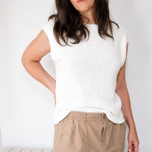 May include: A white sleeveless knit top with a round neckline and a straight hemline, paired with tan trousers featuring an elasticized waistband. The top is constructed from a fine-gauge knit fabric with a clean, simple design and a relaxed fit.