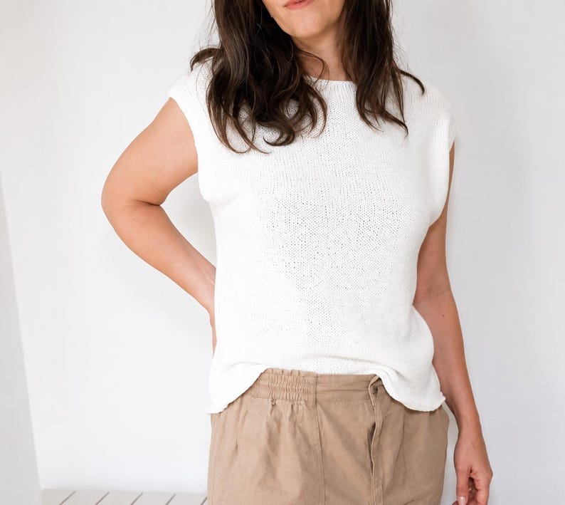 May include: A white sleeveless knit top with a round neckline and a straight hemline, paired with tan trousers featuring an elasticized waistband. The top is constructed from a fine-gauge knit fabric with a clean, simple design and a relaxed fit.