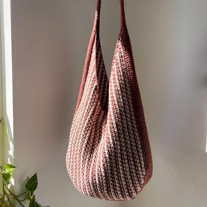 May include: A handmade, open-weave tote bag in a vertical striped pattern of dusty rose and cream. The bag has long, matching straps and a rounded bottom. The bag is held up by a hand.