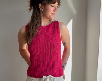 May include: A sleeveless, ribbed knit top in a vibrant magenta colour featuring a high, rounded neckline. The garment is paired with off-white, high-waisted trousers that include a matching fabric tie at the waist, creating a clean, simple silhouette.