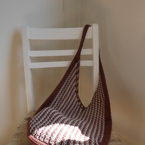 May include: A brown and white striped crochet bag with a long strap, resting on a wooden surface. The bag has a rounded bottom and a wide opening. The bag is sitting on a white chair.