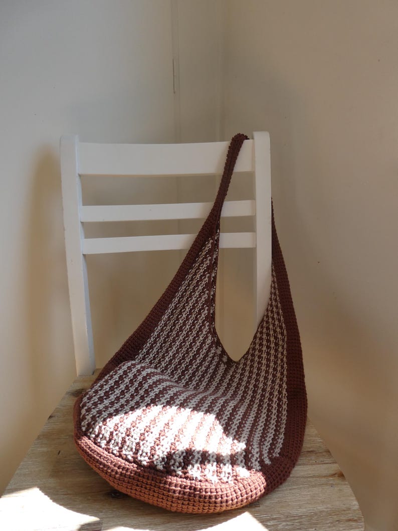 May include: A brown and white striped crochet bag with a long strap, resting on a wooden surface. The bag has a rounded bottom and a wide opening. The bag is sitting on a white chair.