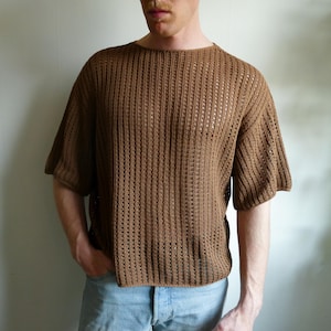 May include: A brown, short-sleeved knit shirt with a textured, open weave design. The shirt has a crew neck and a relaxed fit. The color is a warm, earthy brown. The shirt is made of a lightweight material.