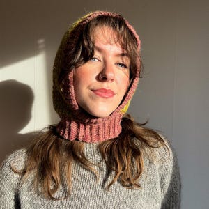 May include: A handmade crochet hood in shades of pink and yellow, worn over a gray knit sweater. The hood frames the face and neck, offering warmth and a unique style. The hood's texture and color contrast with the sweater, creating a cozy, textured look.