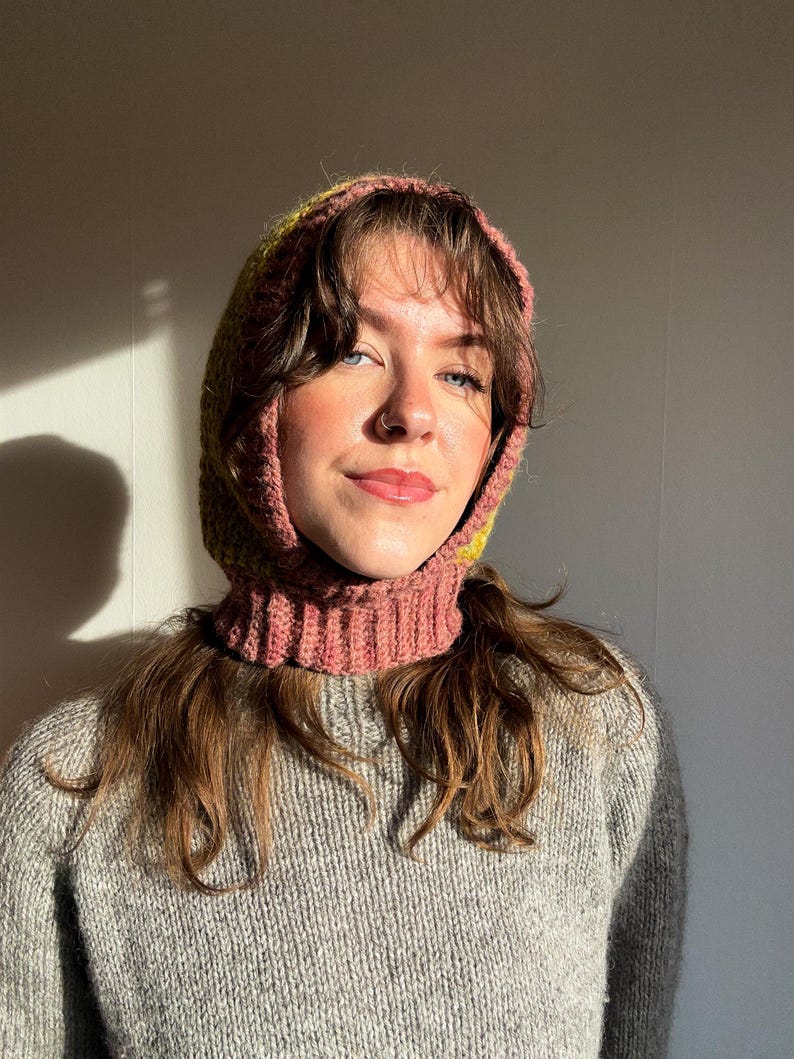 May include: A handmade crochet hood in shades of pink and yellow, worn over a gray knit sweater. The hood frames the face and neck, offering warmth and a unique style. The hood's texture and color contrast with the sweater, creating a cozy, textured look.