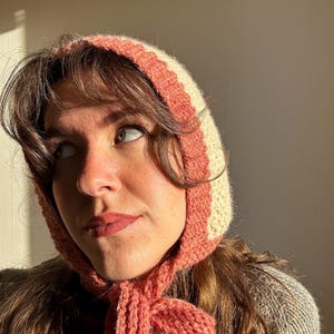 May include: A knitted bonnet in cream and coral. The bonnet has a striped design and ties under the chin. The person in the image is wearing a gray sweater. The bonnet is a vintage style.