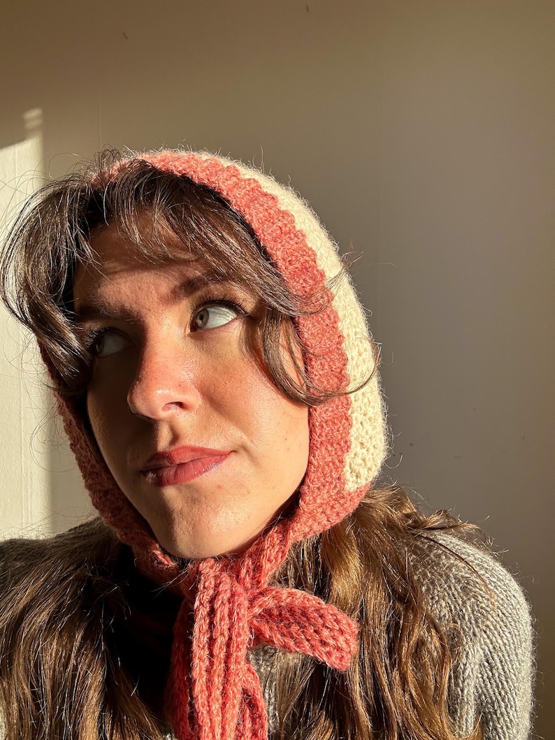 May include: A knitted bonnet in cream and coral. The bonnet has a striped design and ties under the chin. The person in the image is wearing a gray sweater. The bonnet is a vintage style.